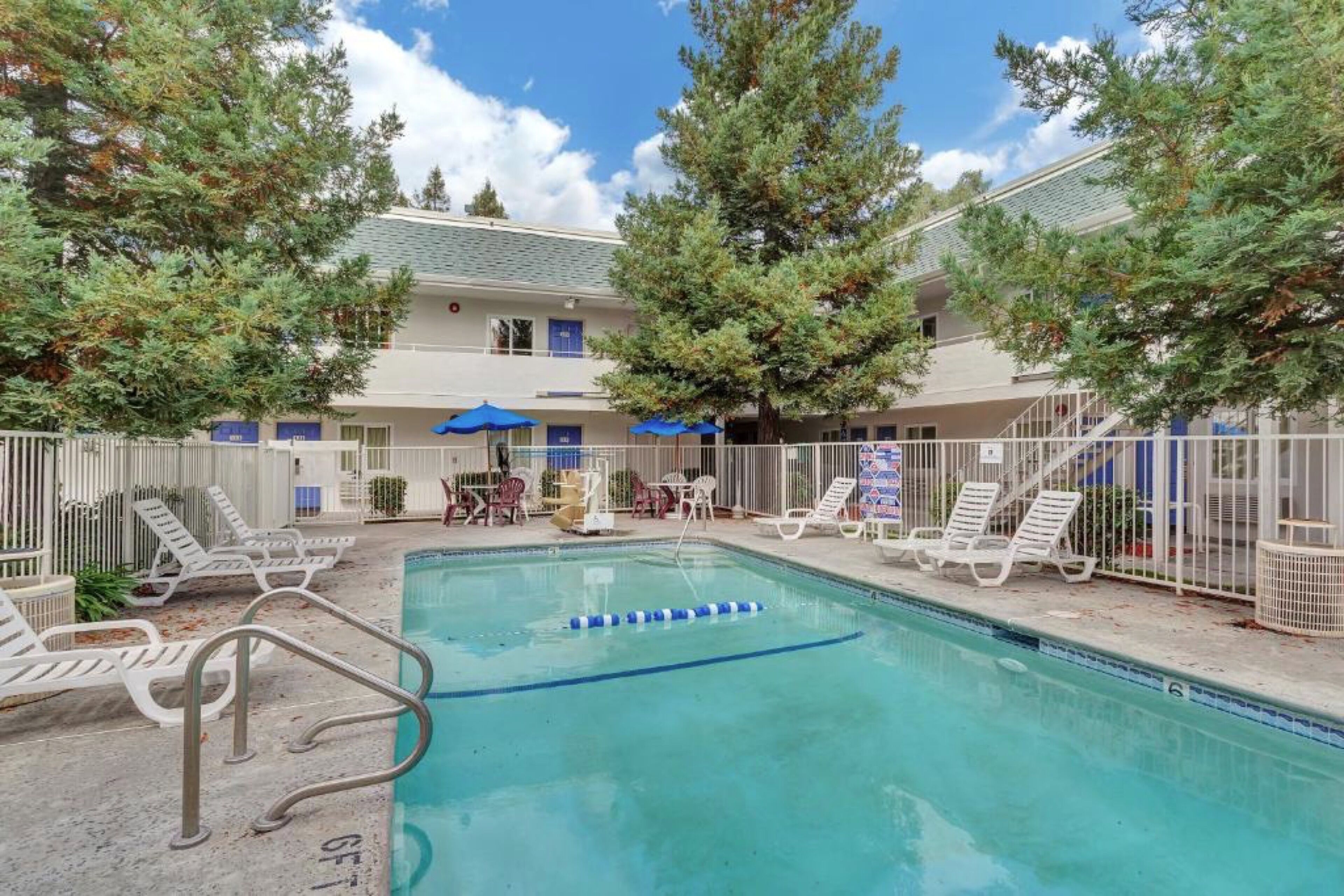 A swimming pool with lounge chairs and umbrellas, surrounded by a fence and trees.