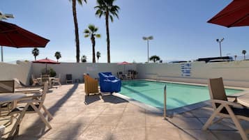 An outdoor swimming pool area with lounge chairs and umbrellas, surrounded by palm trees.