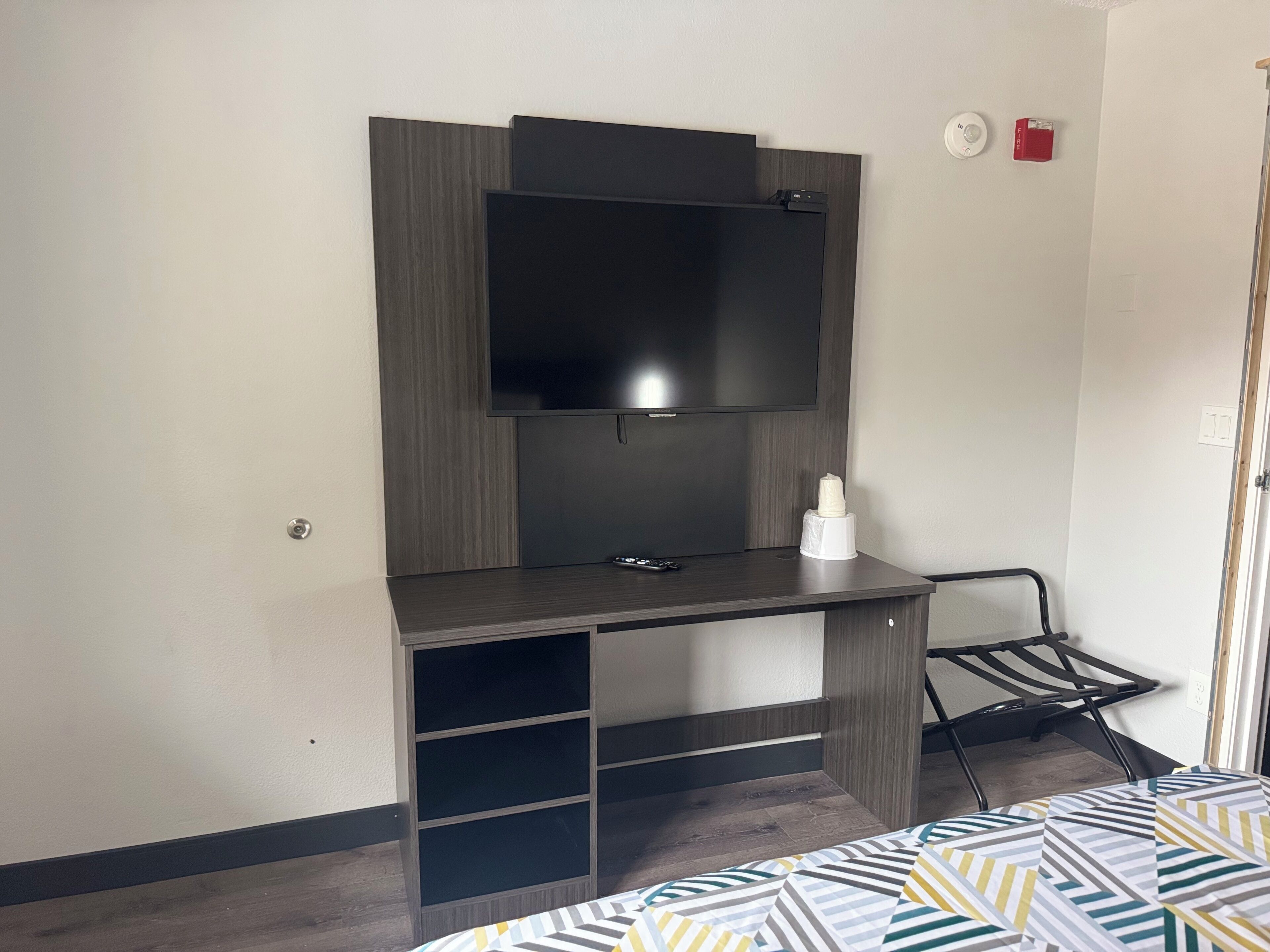 A room with a dark wood desk, a flat-screen TV mounted above it, a small white object on the desk, and a metal rack on the floor.
