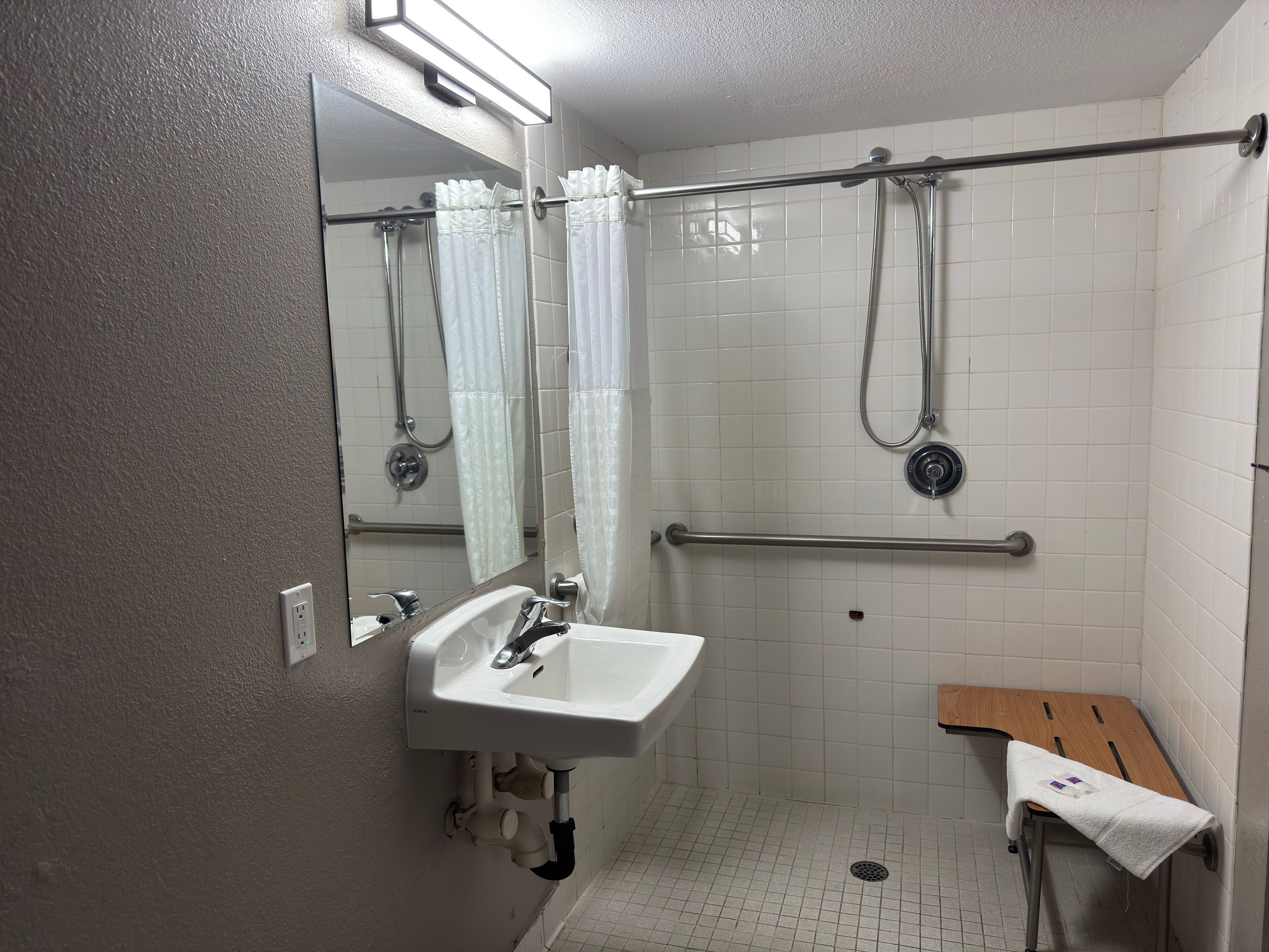 A bathroom with a white sink, a mirror, a shower with a grab bar, and a wooden bench.