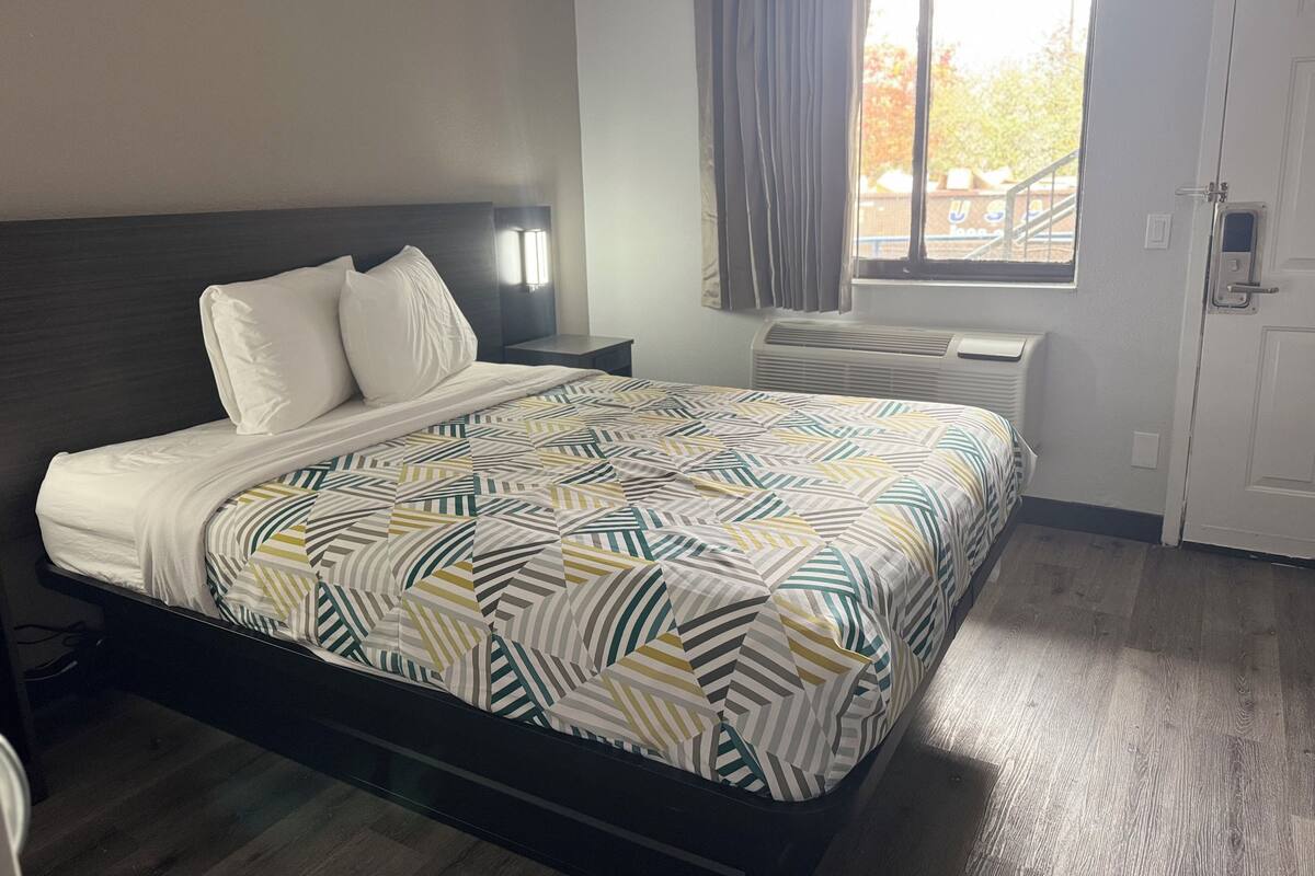 Una habitación de hotel con cama, mesita de noche, ventana con cortinas y equipo de aire acondicionado.