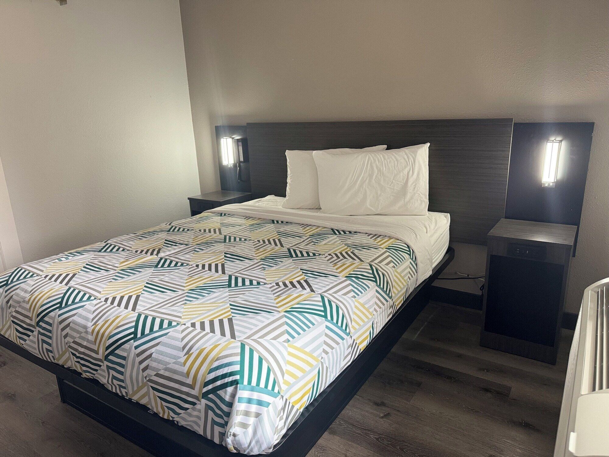 Una cama bien hecha con colcha estampada y almohadas blancas, una mesita de noche con lámpara y un cabecero oscuro.