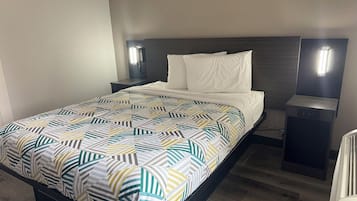 Una cama bien hecha con colcha estampada y almohadas blancas, una mesita de noche con lámpara y un cabecero oscuro.