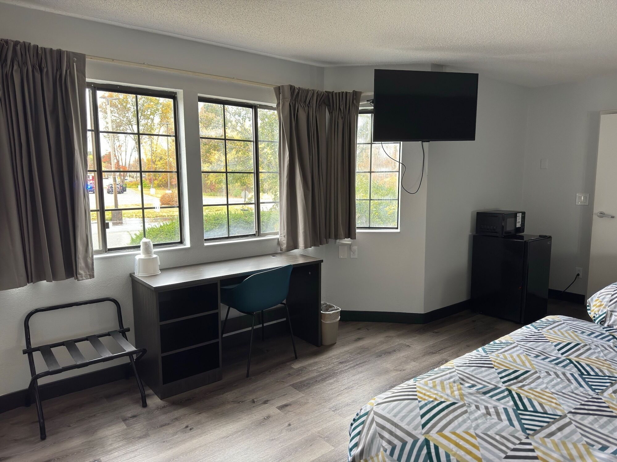 A hotel room with a desk, chair, bed, TV, and windows with curtains.