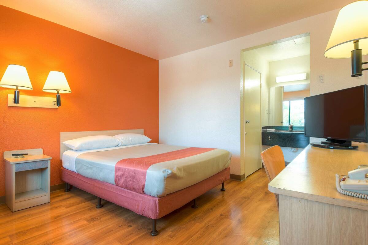 A hotel room with a bed, bedside tables, a desk with a chair, a television, and a bathroom visible through an open door.