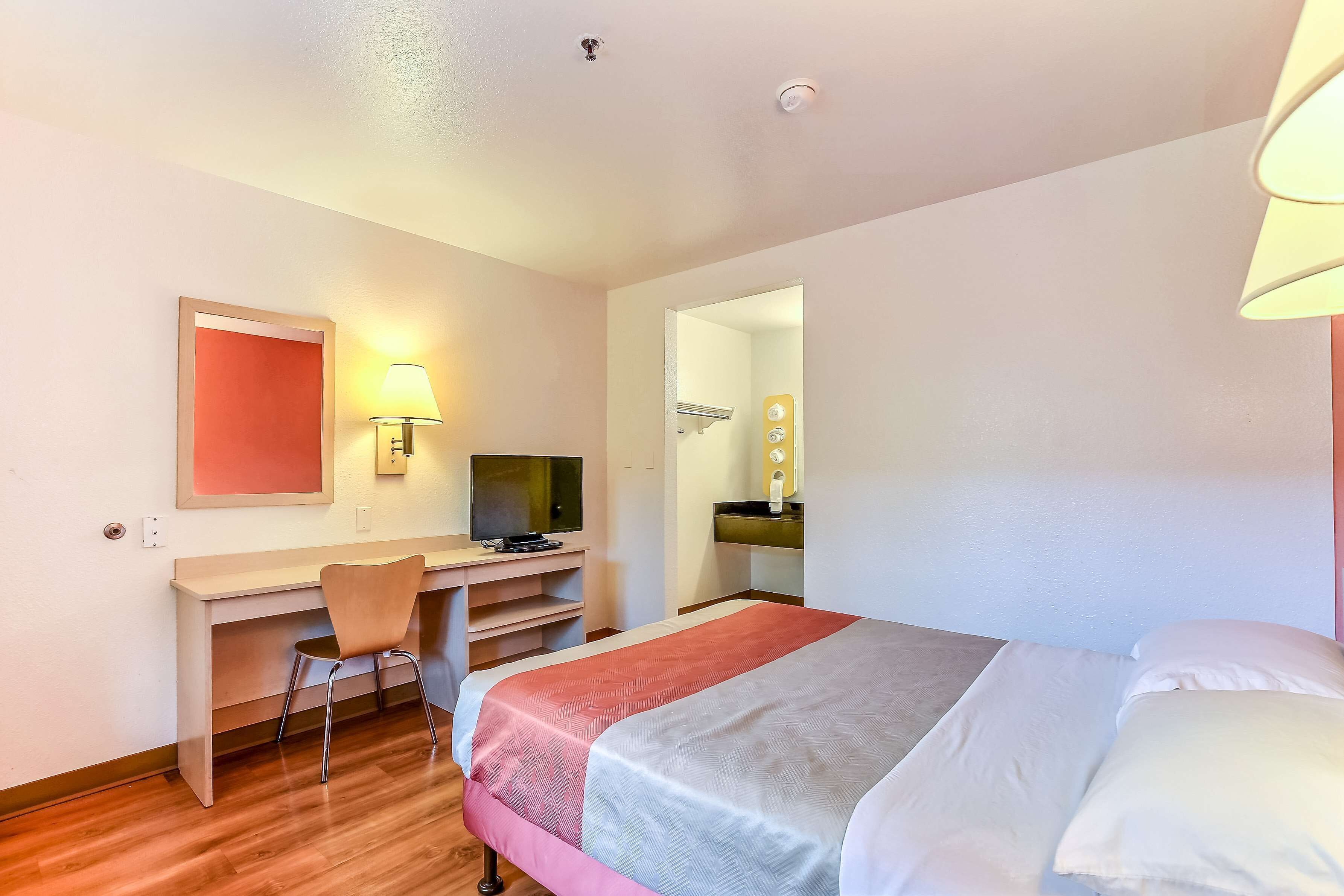 A hotel room with a bed, a desk, a chair, a television, and a bathroom visible through an open door.