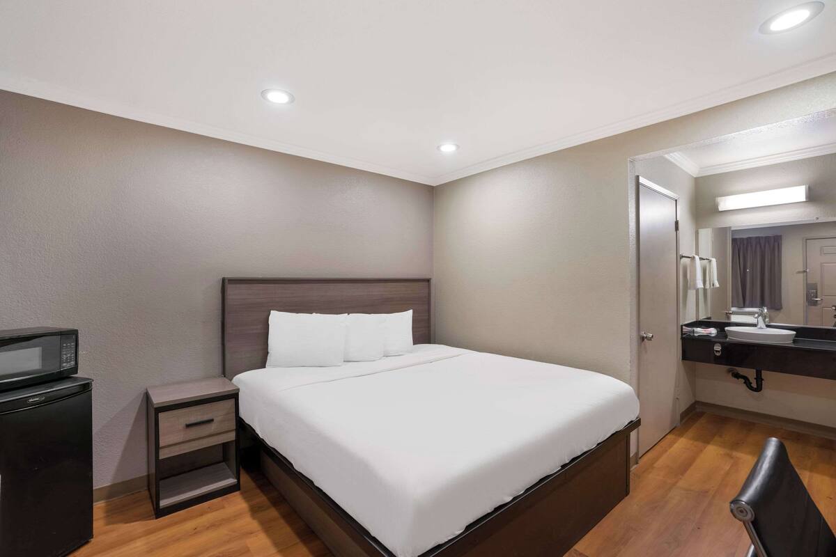 A hotel room with a large bed, a nightstand with a microwave, a chair, and a bathroom with a sink and mirror.