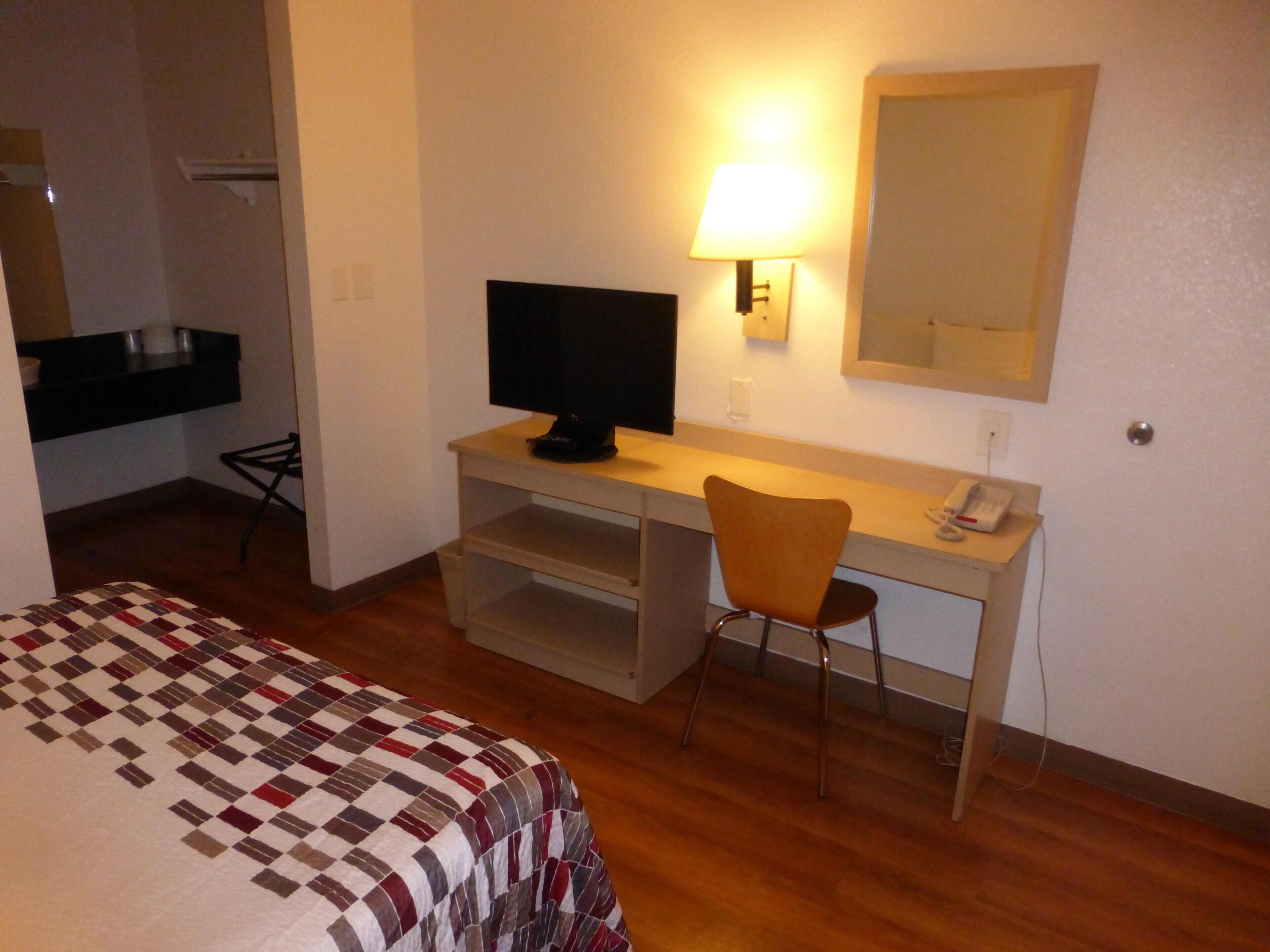 A hotel room with a bed, a desk with a television, a chair, a desk lamp, a telephone, and a mirror.