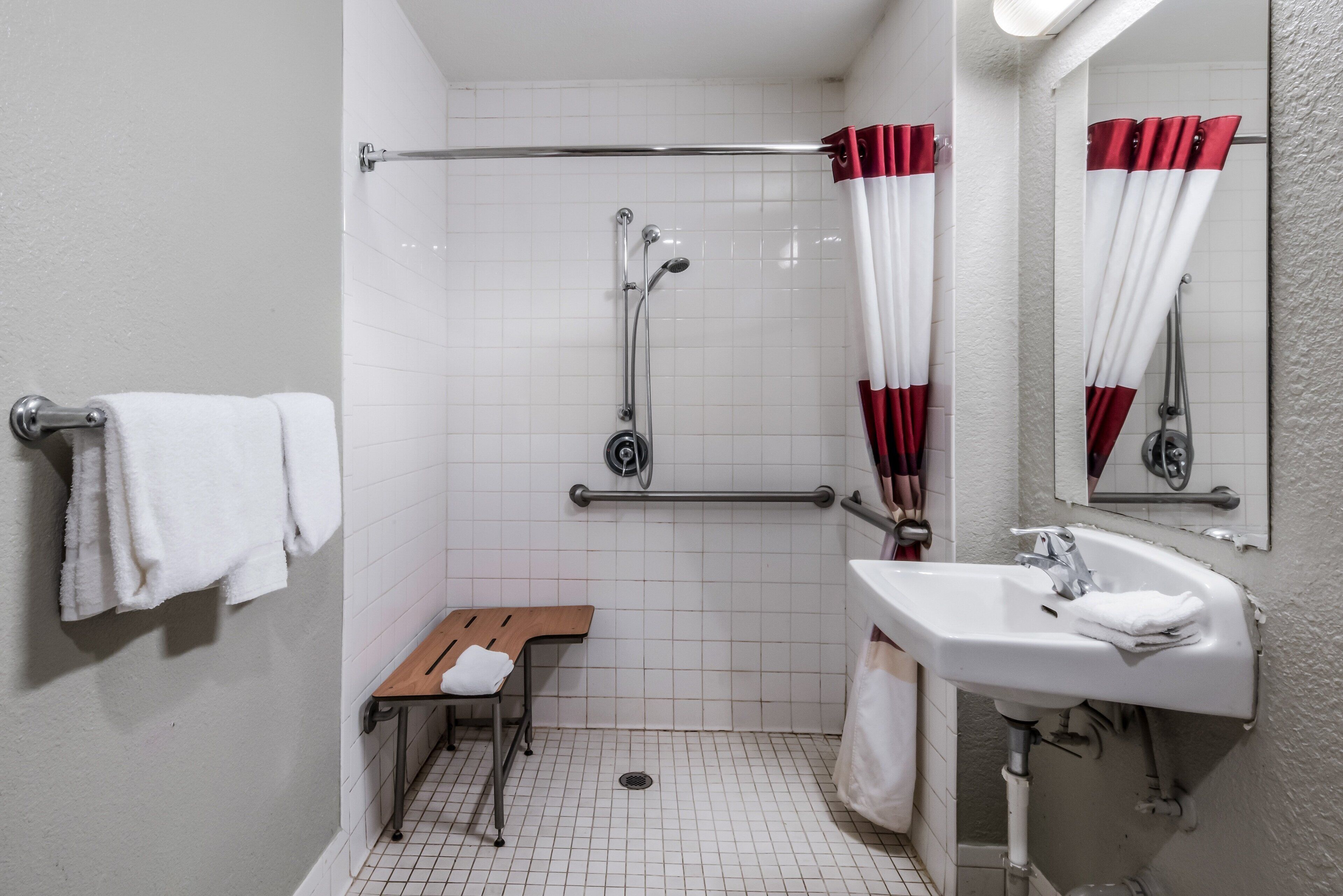 Une salle de bain avec un lavabo blanc, un miroir, une douche équipée d’une barre d’appui, un banc et un porte-serviettes.