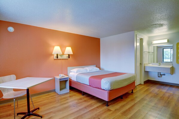 Una habitación de hotel con cama, mesita de noche, lámpara, silla y baño con lavabo y espejo.