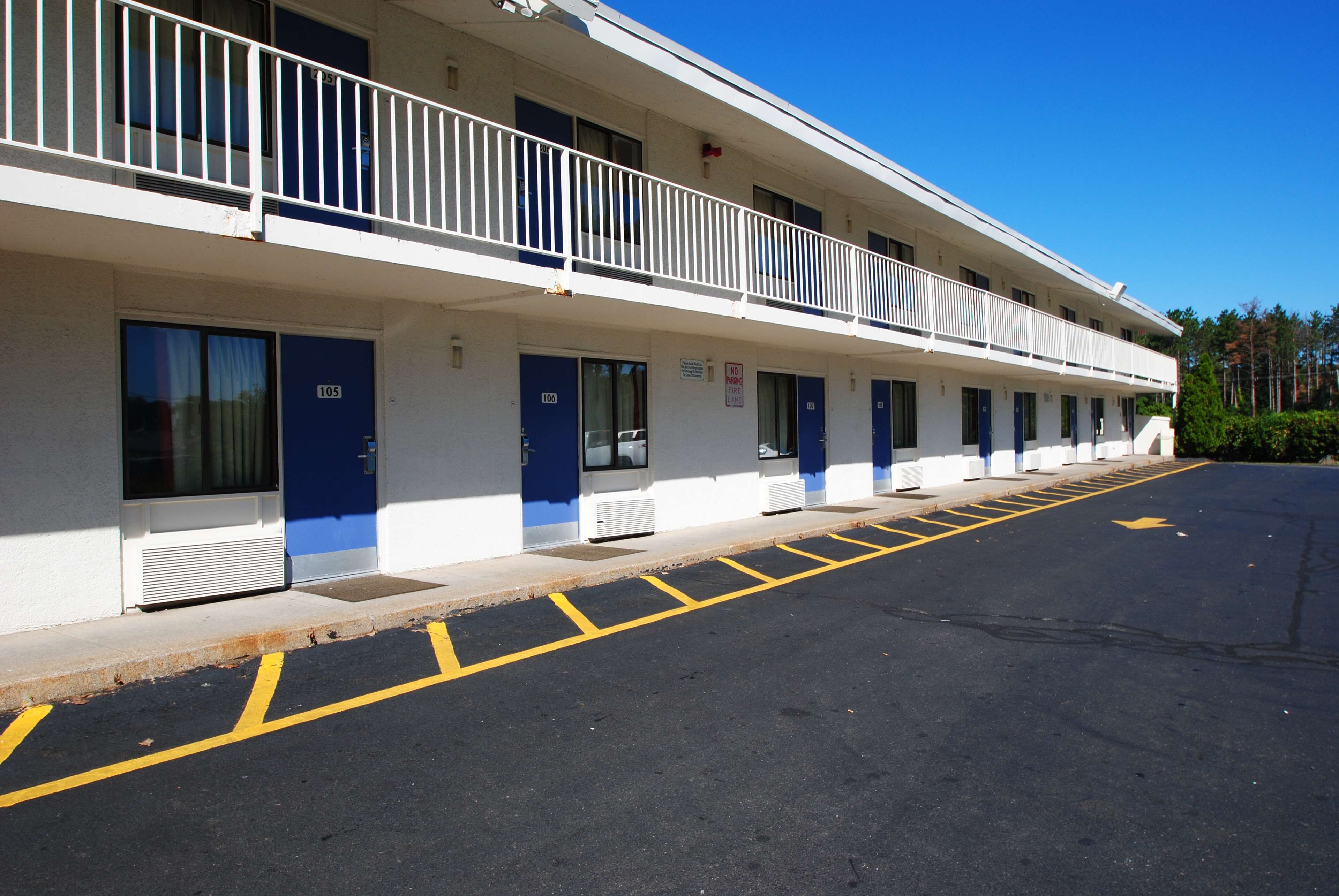A motel with a row of rooms, each with a blue door and a number. There is a parking lot with marked spaces and a no parking sign.
