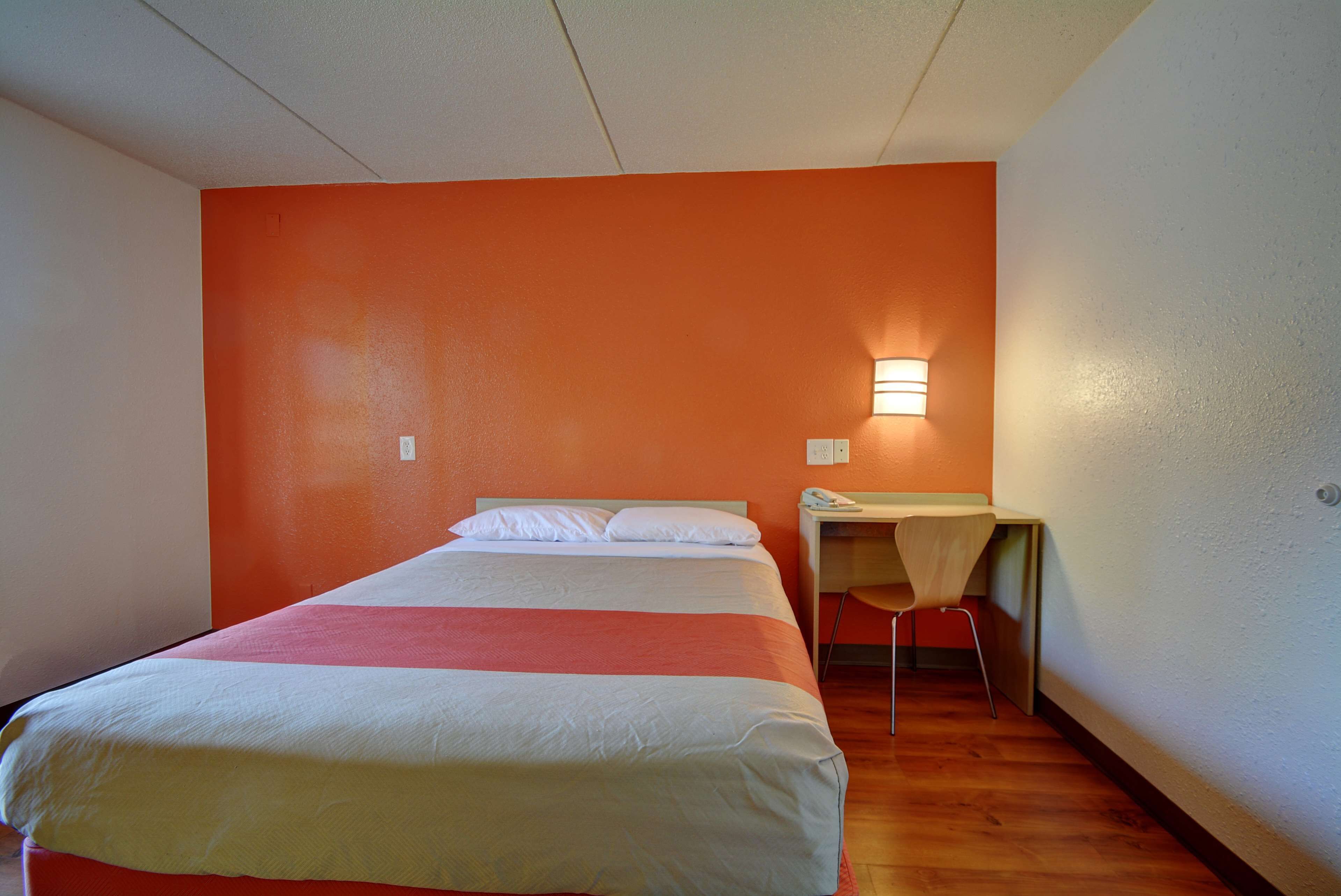 Een eenpersoonskamer met een oranje accentmuur, een bureau met stoel, en een wandlamp.