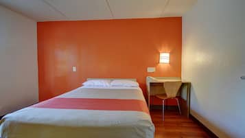 Quarto individual com uma parede de destaque laranja, uma escrivaninha com cadeira e uma luminária fixada na parede.