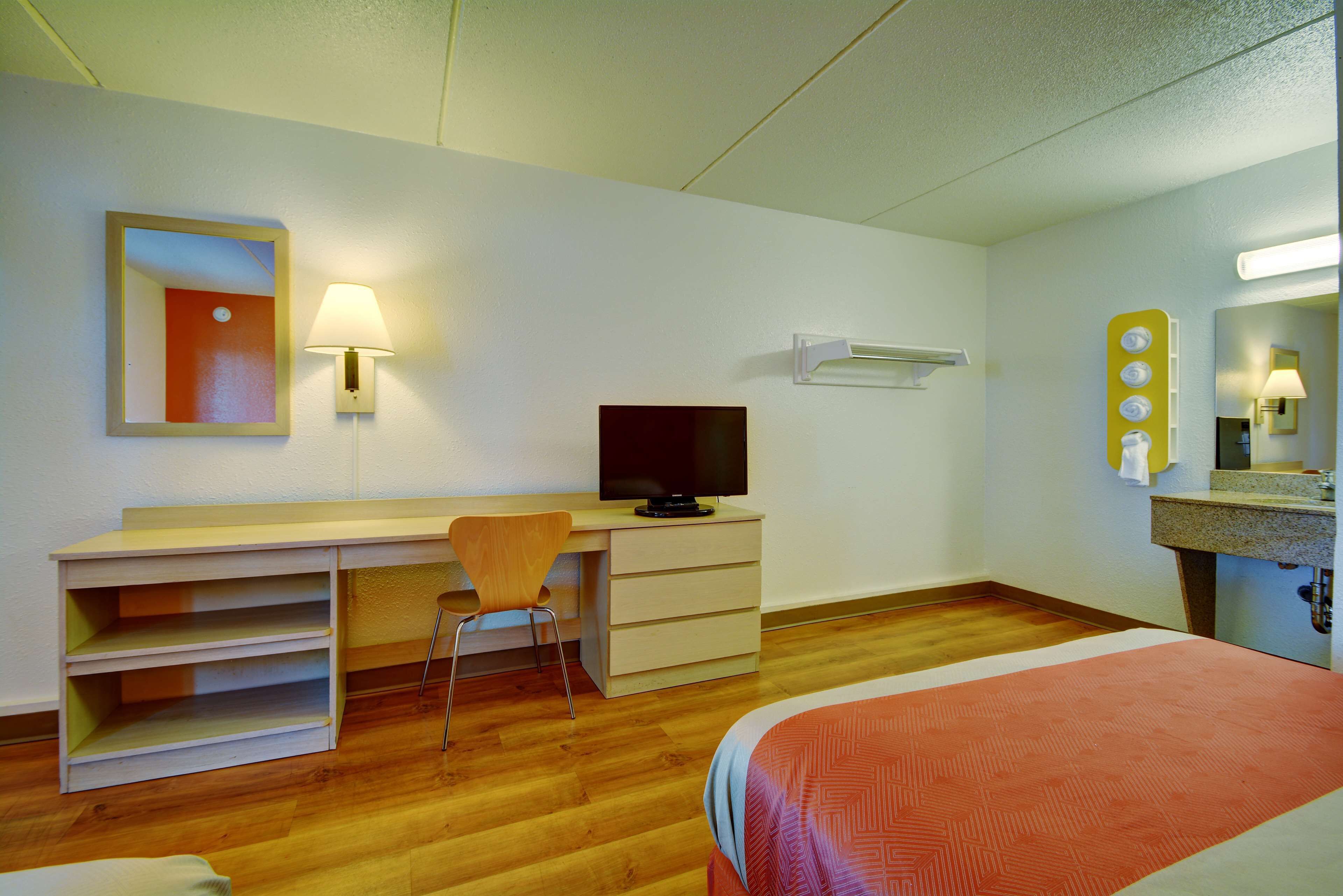 A hotel room with a wooden desk, a television, a chair, a bed with a patterned bedspread, a mirror, and a wall-mounted lamp.