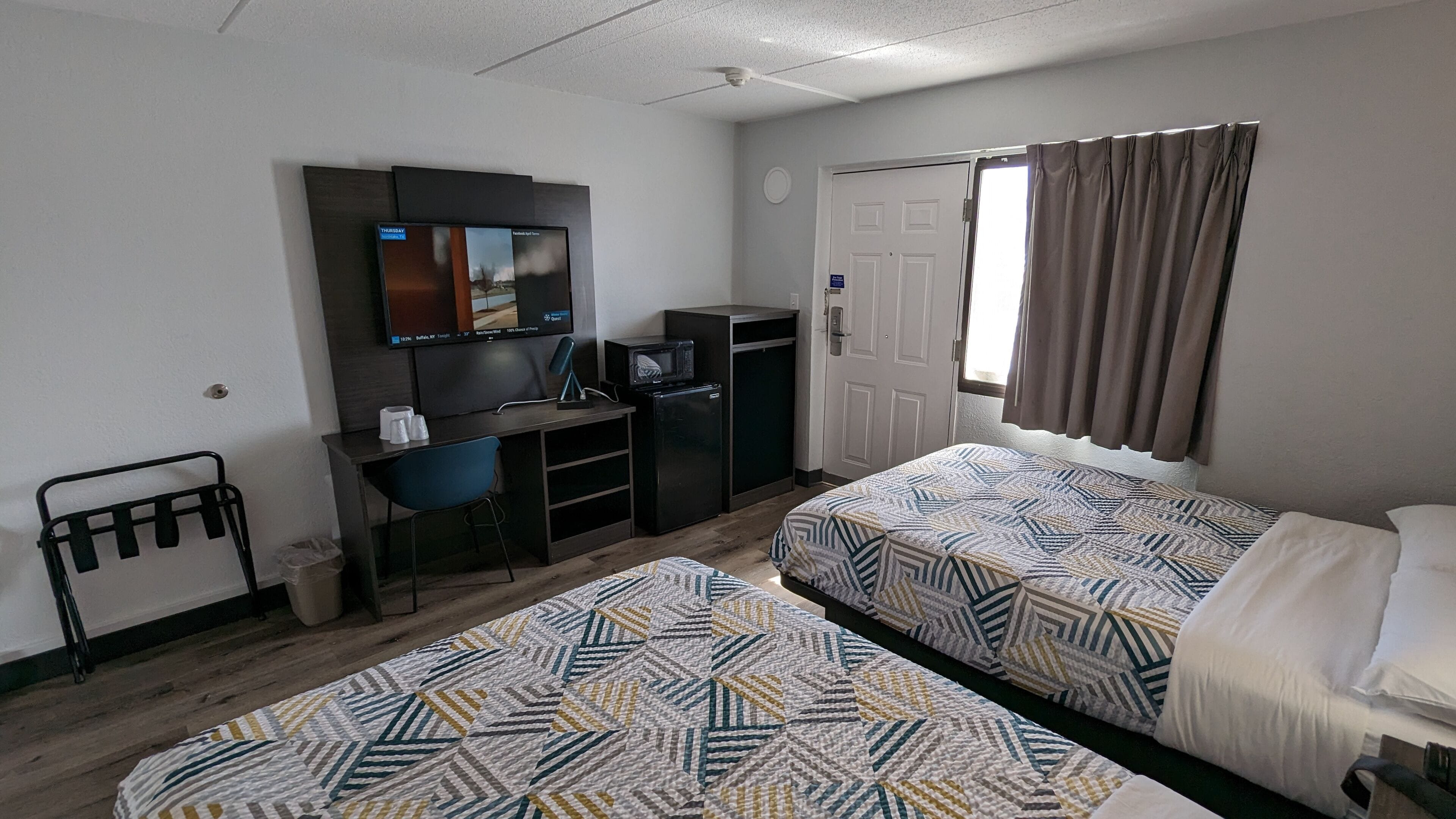 A hotel room with two beds, a desk with a television, a small refrigerator, and a window with curtains.