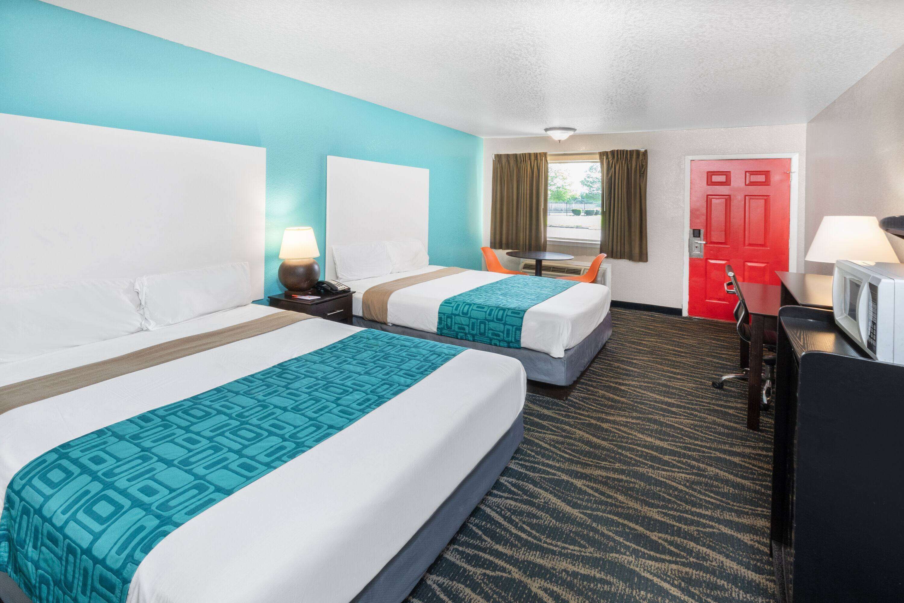 A hotel room with two beds, a desk, a chair, a TV, and a red door.