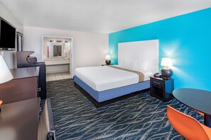 A hotel room with a bed, a desk, a chair, a TV, and a bathroom with a mirror and sink.