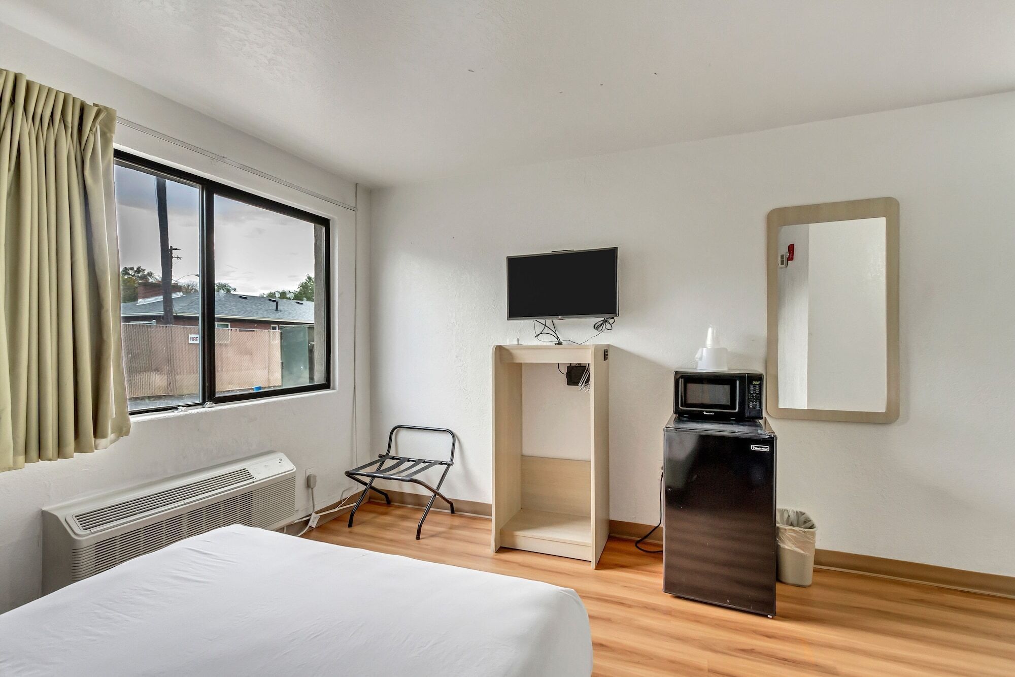 A hotel room with a bed, a desk with a computer and microwave, a TV, a chair, and a window with curtains.