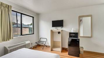 A hotel room with a bed, a desk with a computer and microwave, a TV, a chair, and a window with curtains.