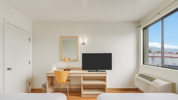 Habitación de hotel con cama, escritorio con computadora, una silla, un espejo y ventana con vista a edificios.