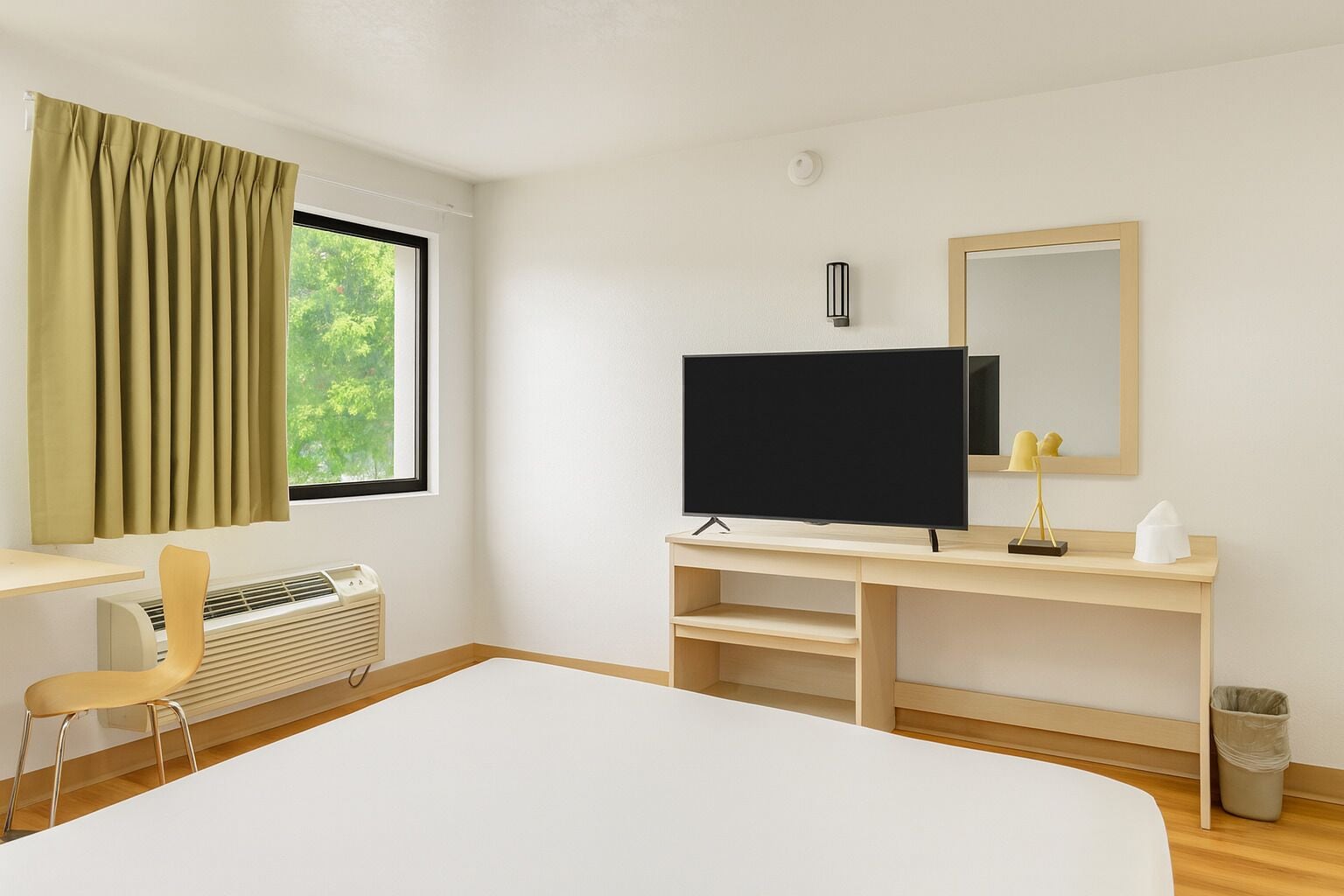Una habitación de hotel moderna con una cama grande, un escritorio con silla, un televisor de pantalla plana, un espejo y un ventanal con cortinas.