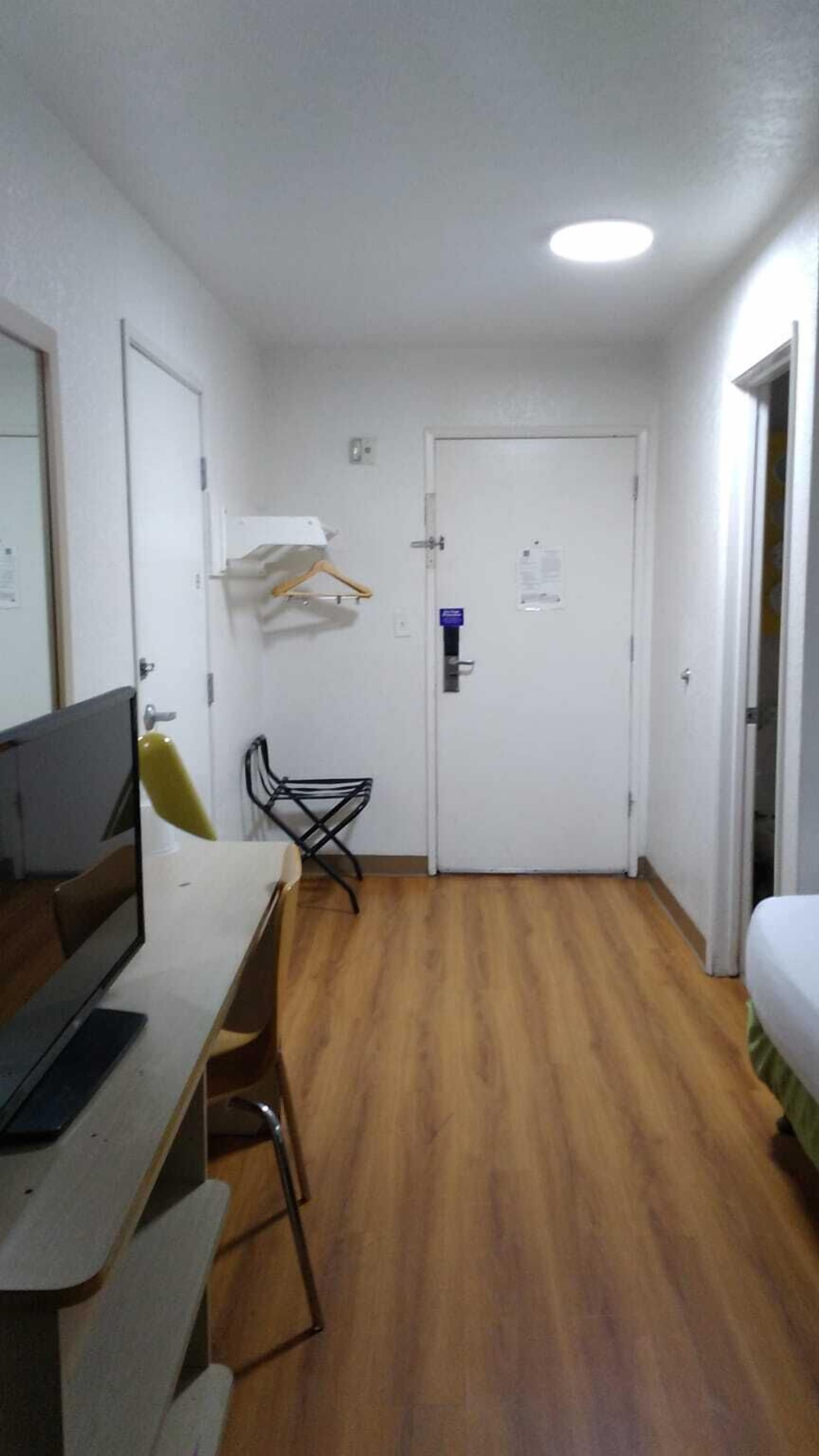 A small, compact room with a wooden floor, a desk with a computer, a chair, a bed, and a door with a lock.