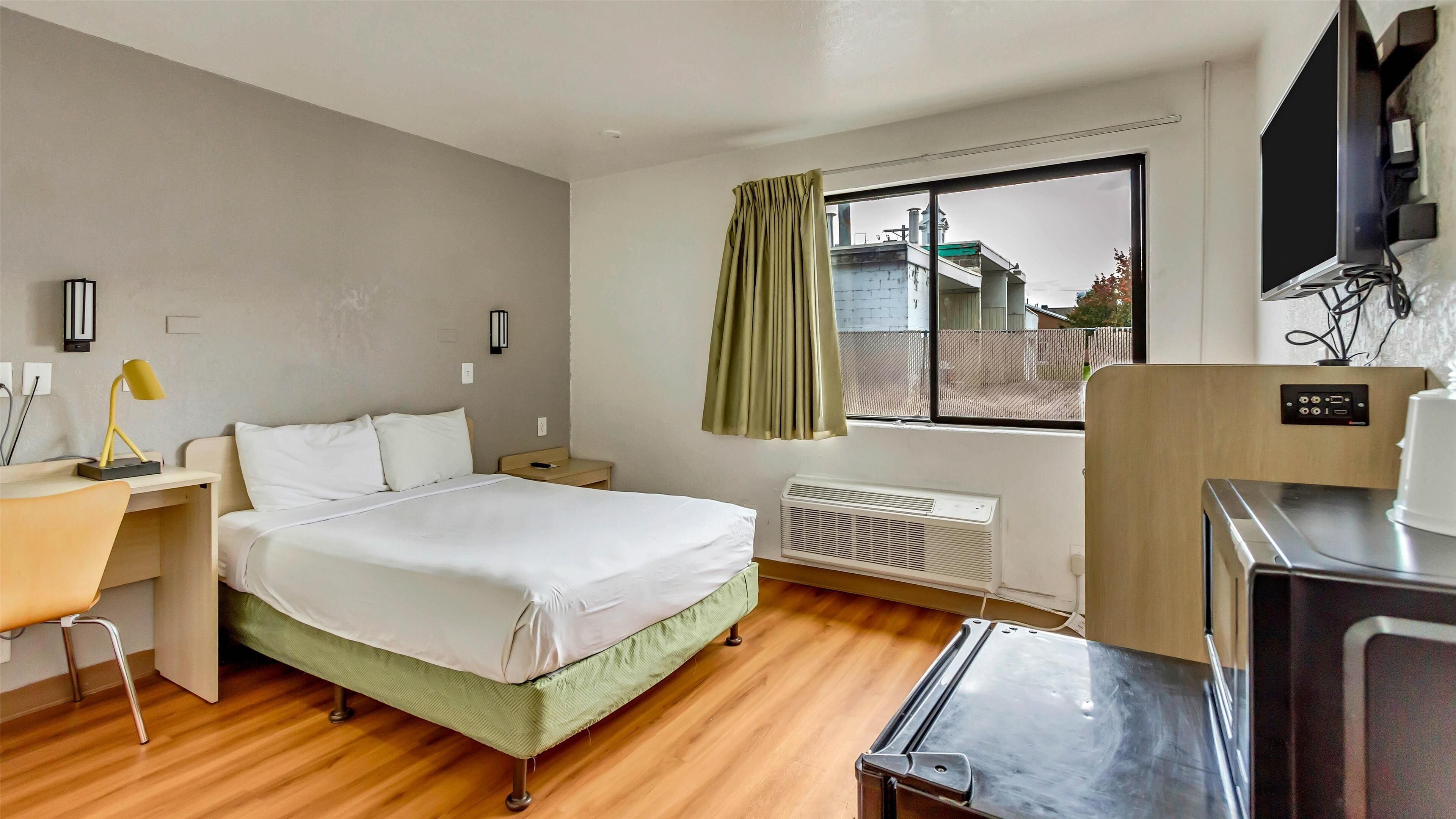 Habitación de hotel con cama, escritorio, silla, ventana con cortinas, radiador y televisión.