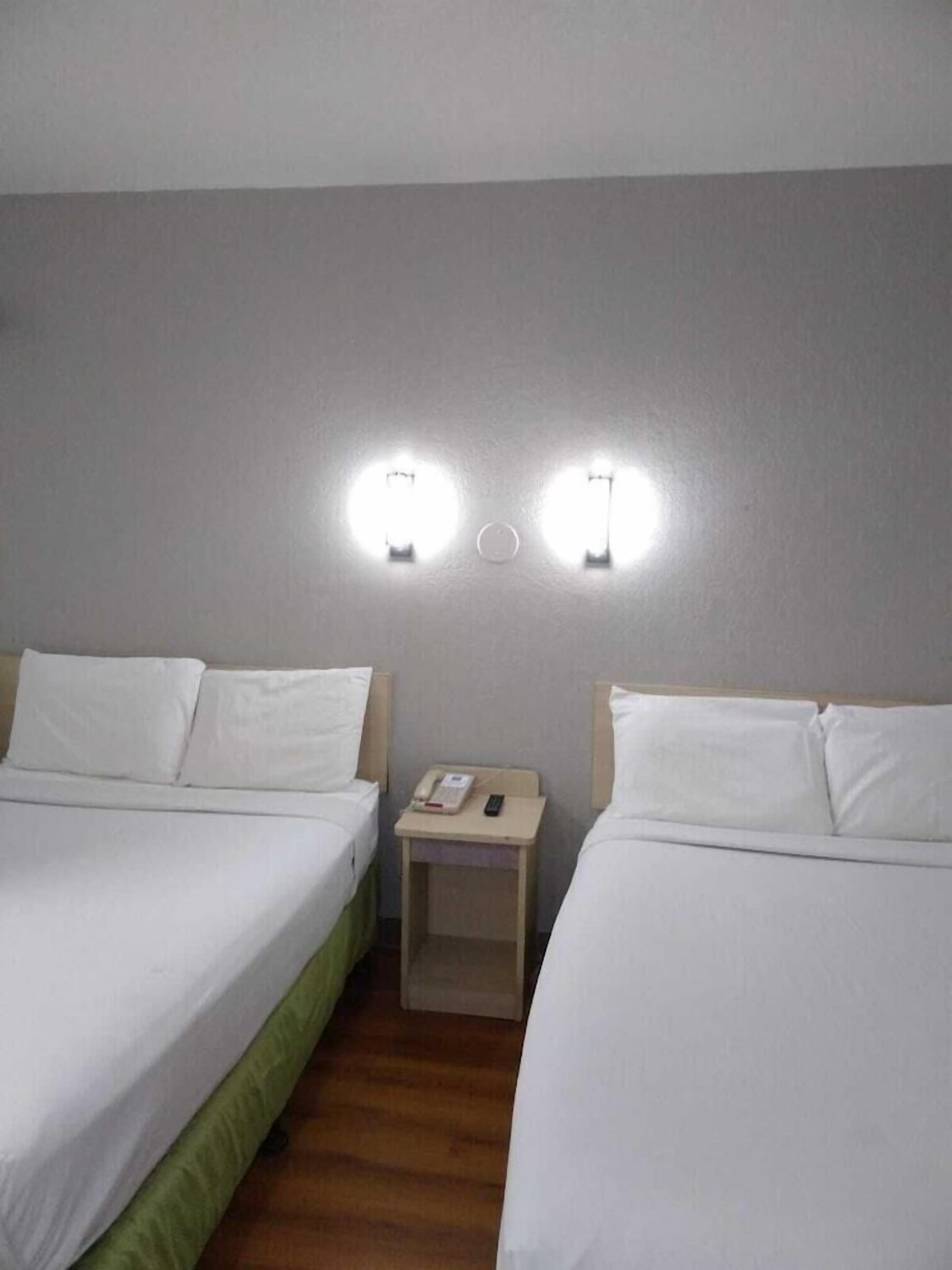Ein Hotelzimmer mit zwei Einzelbetten, einem kleinen Nachttisch mit Telefon und Fernbedienung sowie Wandleuchten.