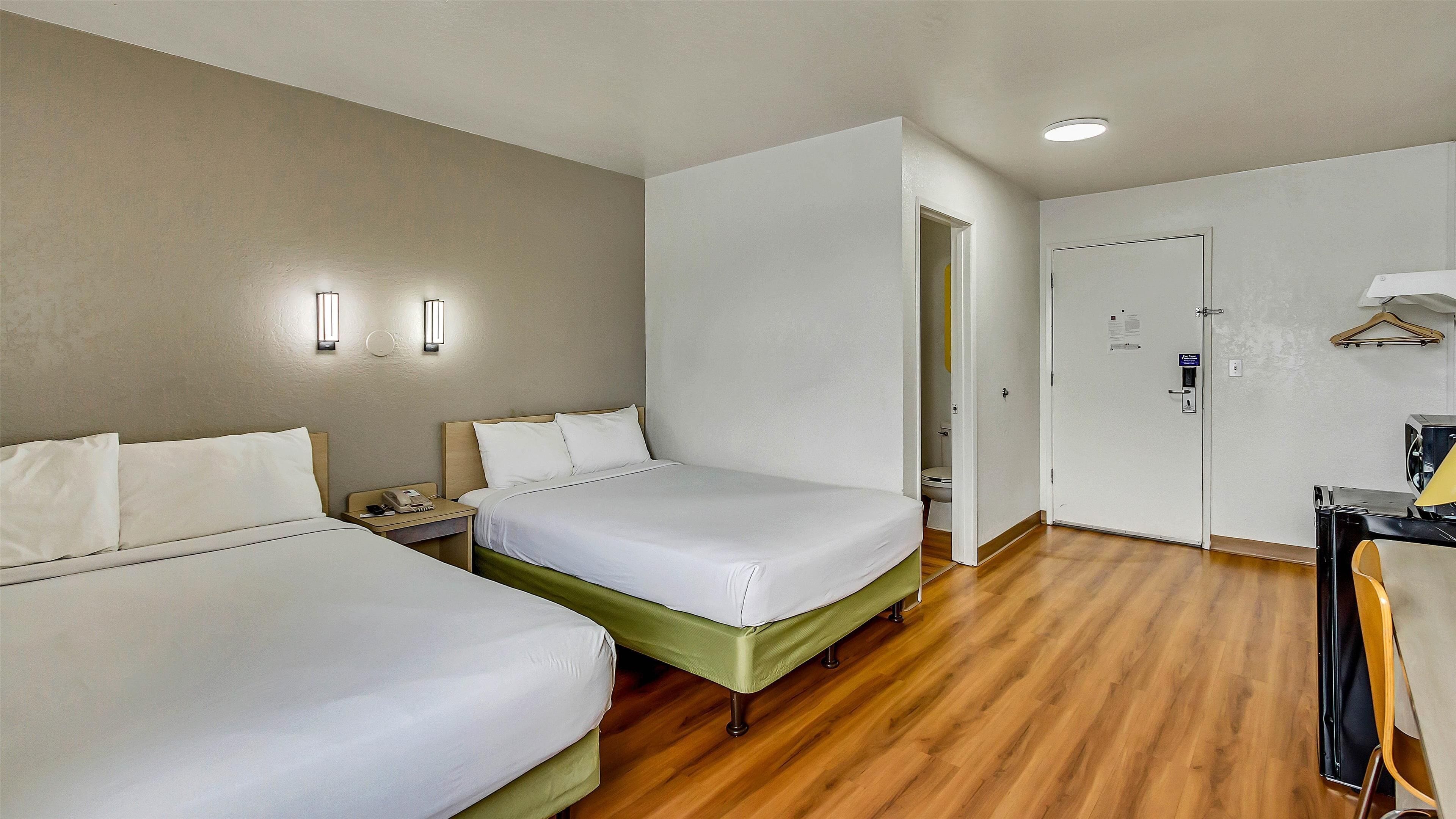 Ein Hotelzimmer mit zwei Betten, Holzboden, Schreibtisch mit Stuhl und einer Tür zu einem Badezimmer.