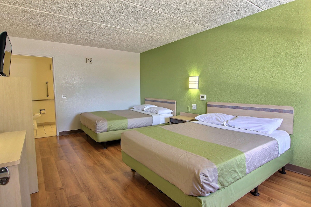 A hotel room with two beds, a wooden floor, a green wall, and a small bedside table with a lamp.