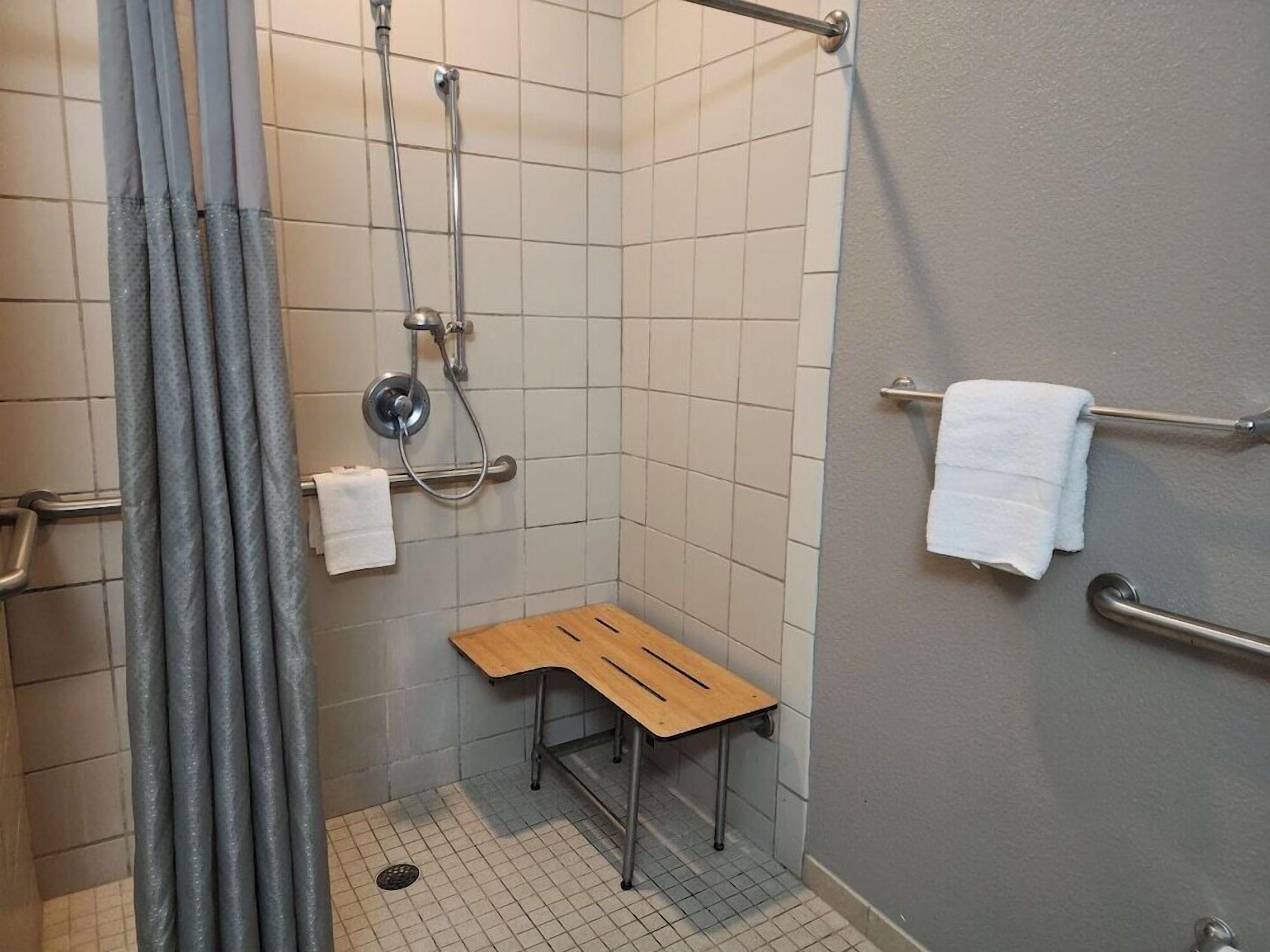 Un petit espace de douche carrelé, avec un banc en bois, des barres d’appui et deux serviettes blanches posées sur un porte-serviettes.