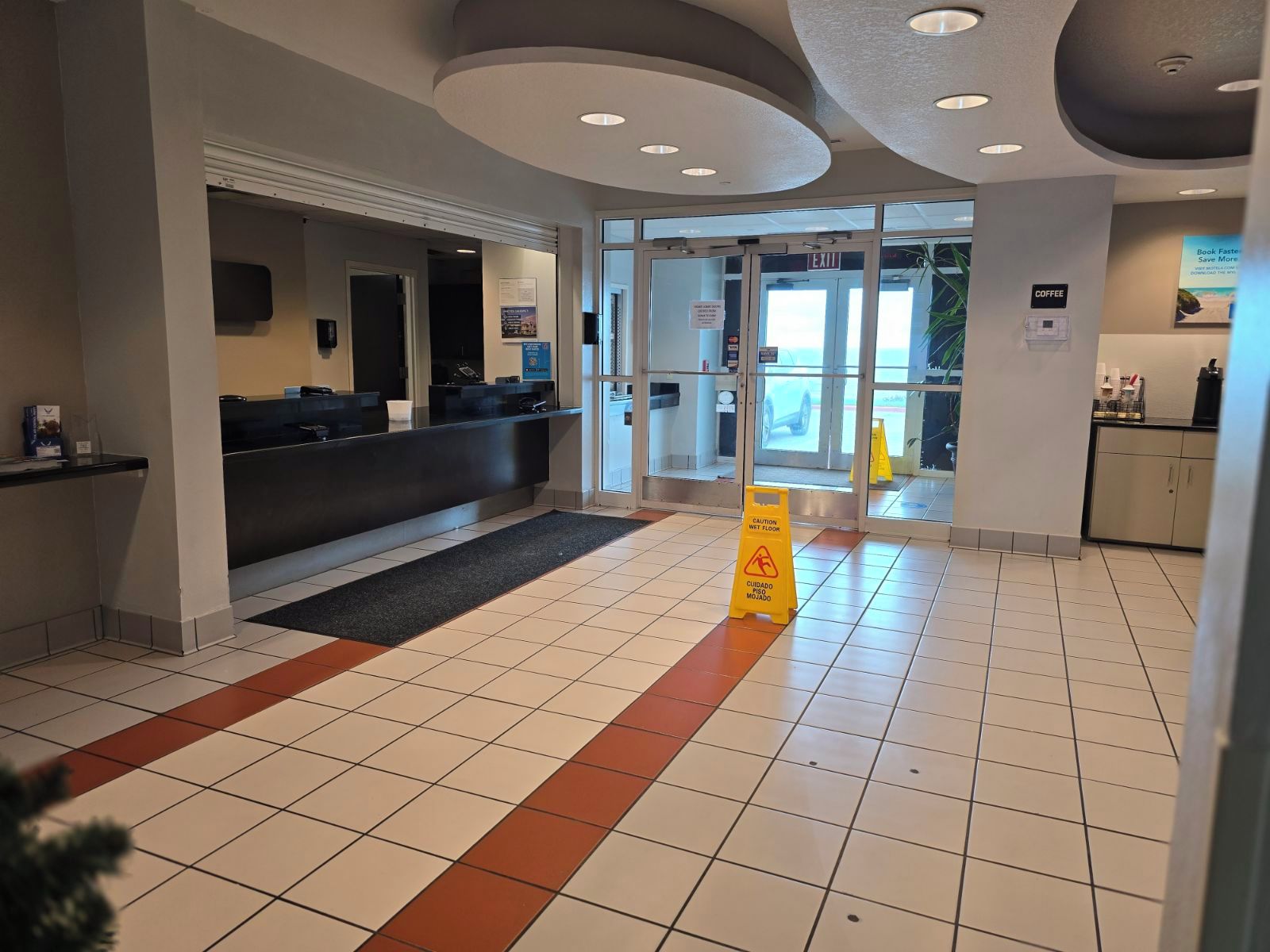 Área de recepção de hotel com balcão de check-in, espaço para café e aviso de chão molhado.