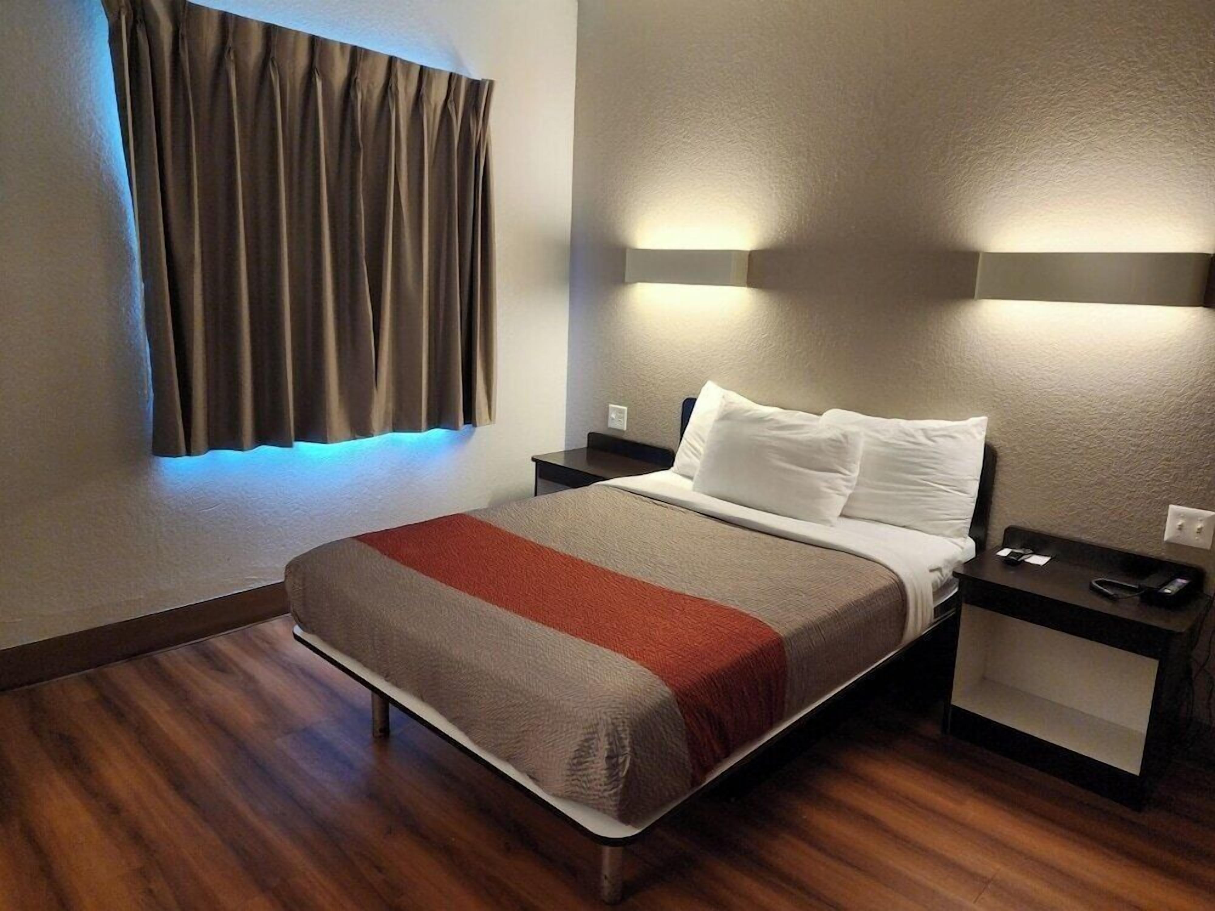 A hotel room with a bed, bedside tables, a nightstand, a window with curtains, and wall-mounted lighting.