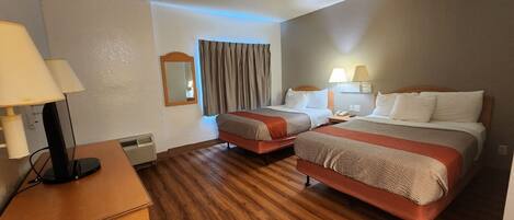A hotel room with two beds, a desk, a lamp, a mirror, and a window with curtains.