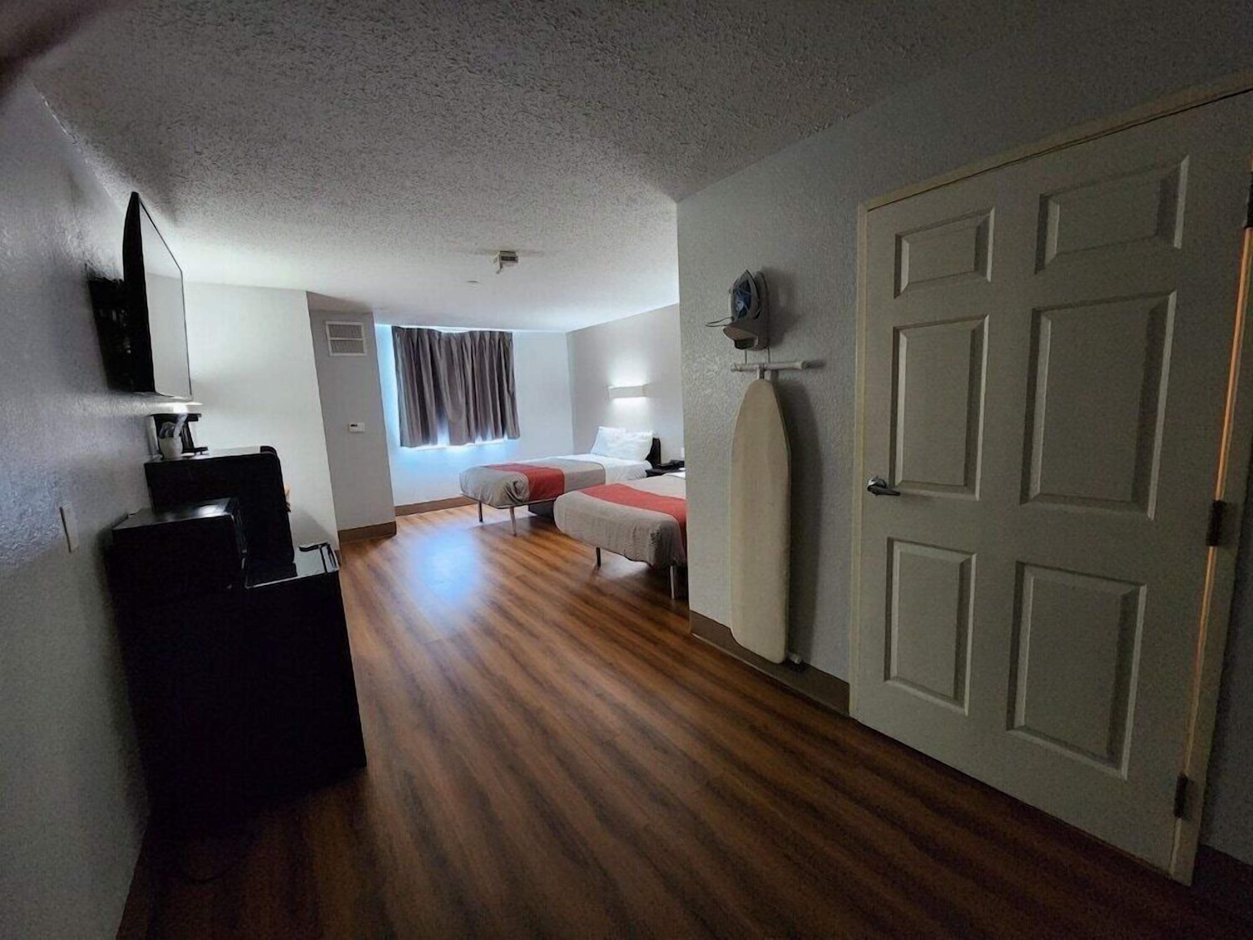 A hotel room with a wooden floor, a bed with a red blanket, a desk with a lamp, and a door with a towel rack.