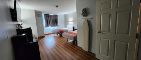 A hotel room with a wooden floor, a bed with a red blanket, a desk with a lamp, and a door with a towel rack.