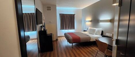 A modern hotel room with a wooden floor, a bed with a red and white bedspread, a desk with a chair, a TV mounted on the wall, and a wardrobe.