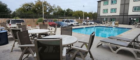 Un área de piscina al aire libre con sillones y mesas, rodeada por una valla y un edificio al fondo.