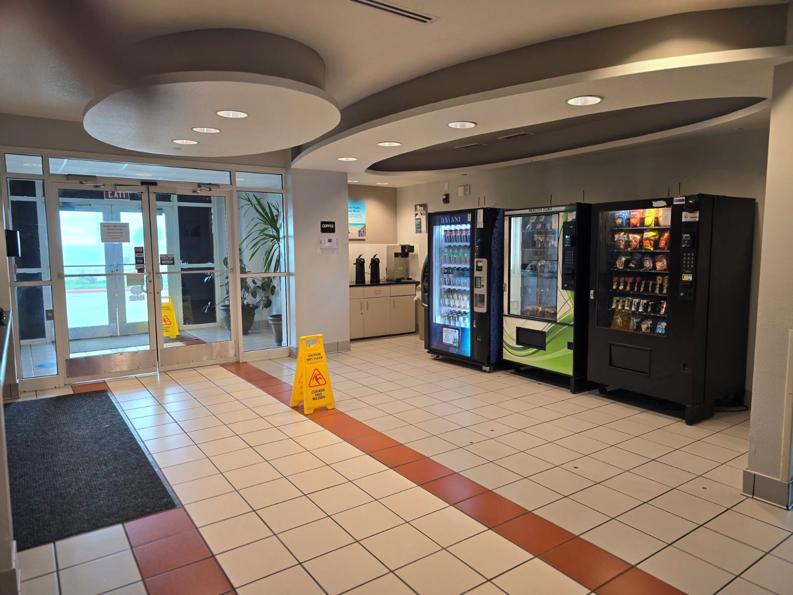A clean, modern interior with a coffee machine, vending machines, and a caution sign on the floor.