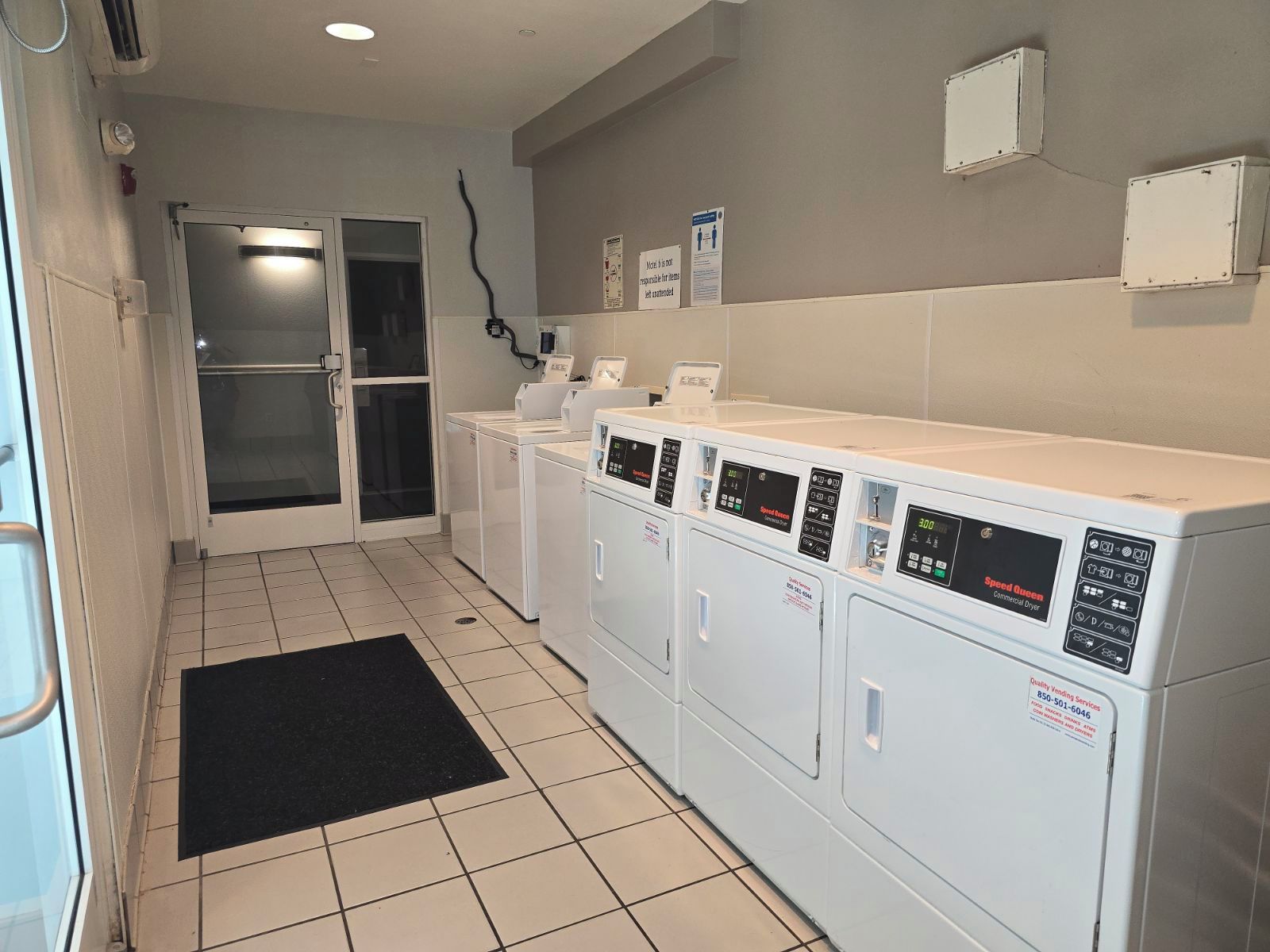 A laundry room with multiple washing machines, a black mat, and a door leading to another area.