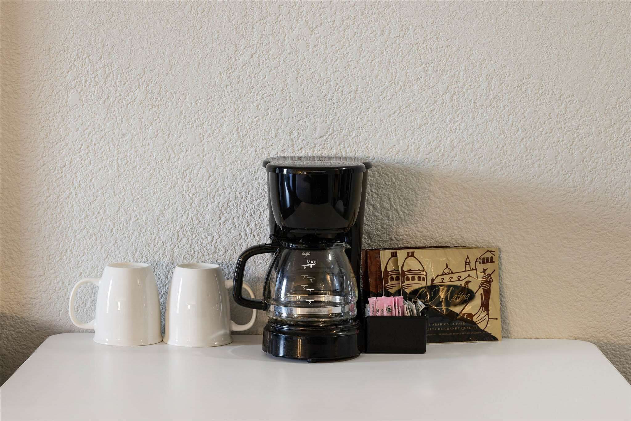 Een zwarte koffiezetapparaat op een witte tafel, met twee witte kopjes ernaast en een koffiezak.