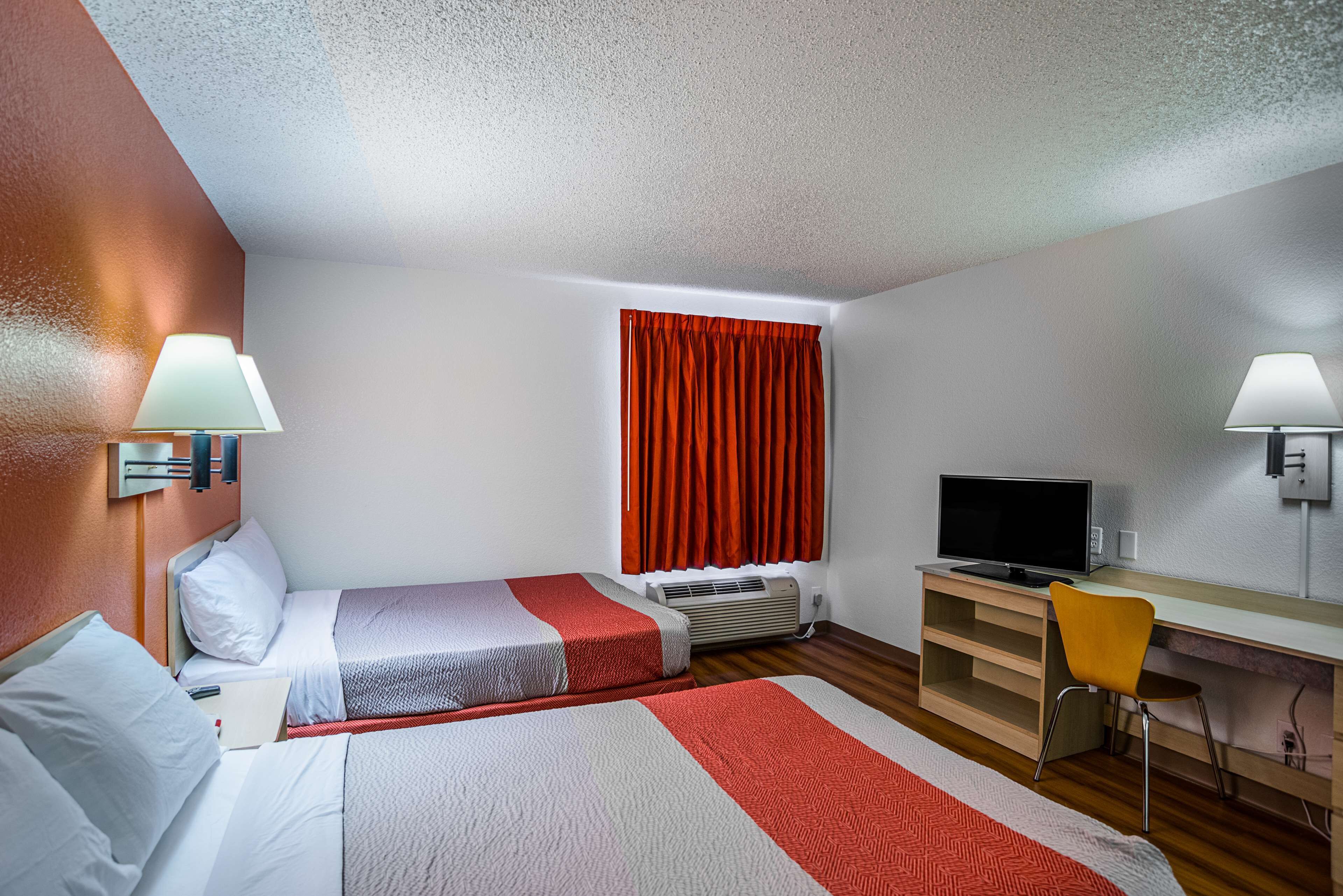 Hotellihuone, jossa on kaksi sänkyä, työpöytä, tuoli, televisio ja ikkuna, jossa on punaiset verhot.