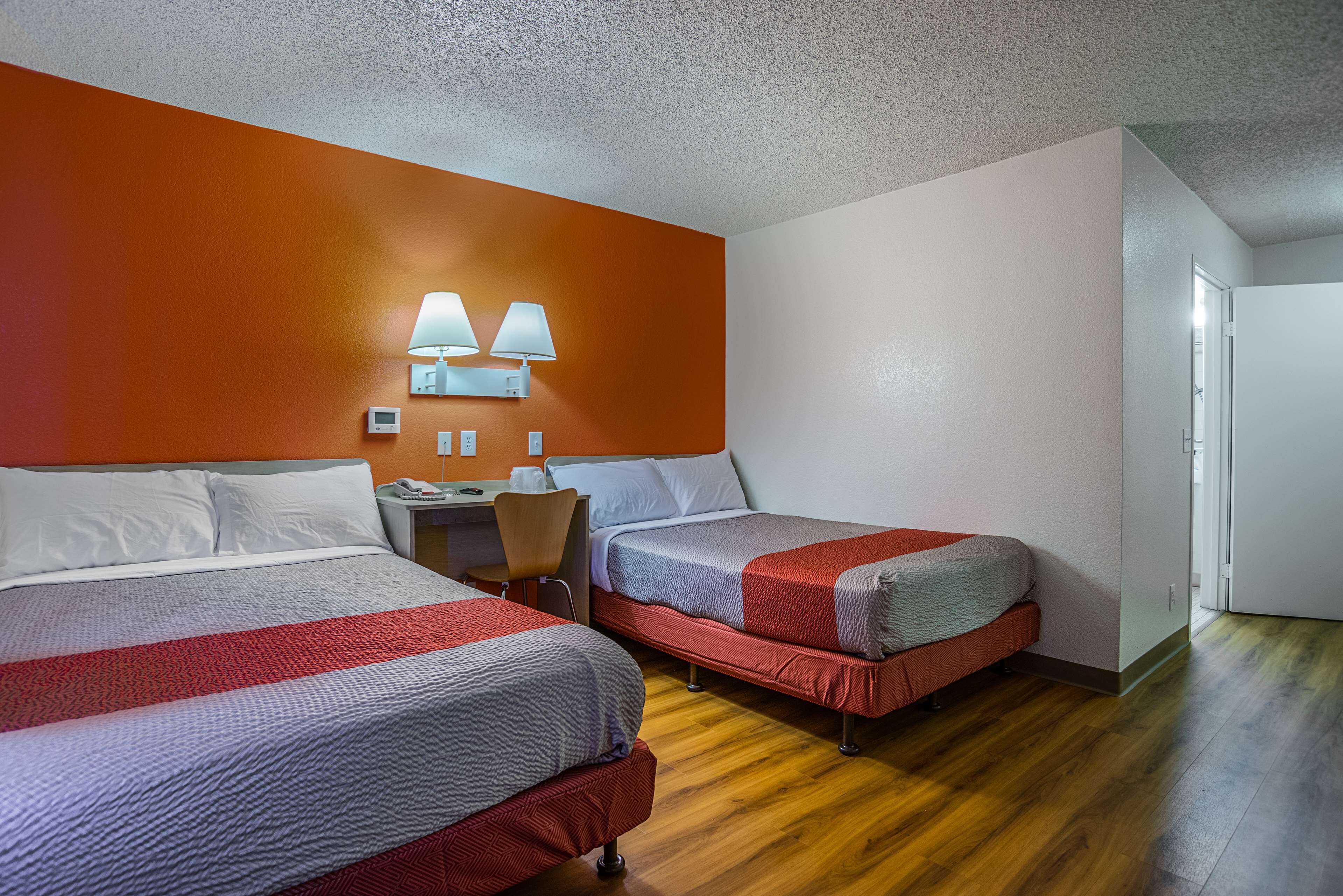 A hotel room with two beds, a desk, and a chair. There are wall-mounted lamps and a thermostat on the wall.