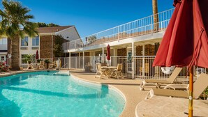 Une piscine avec des chaises longues et des parasols, un espace de détente couvert et un bâtiment doté d’un balcon.