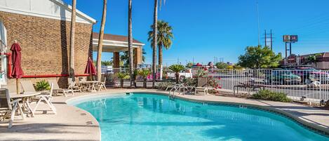 A swimming pool with lounge chairs and umbrellas, surrounded by palm trees and a brick building.