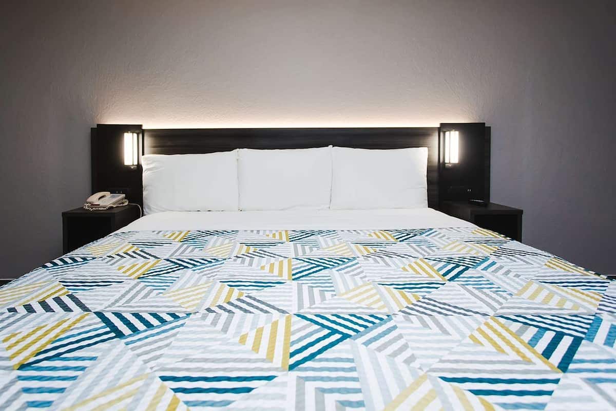 A neatly made bed with a geometric patterned bedspread, a dark headboard with integrated lighting, and a bedside table with a phone and a small decorative item.