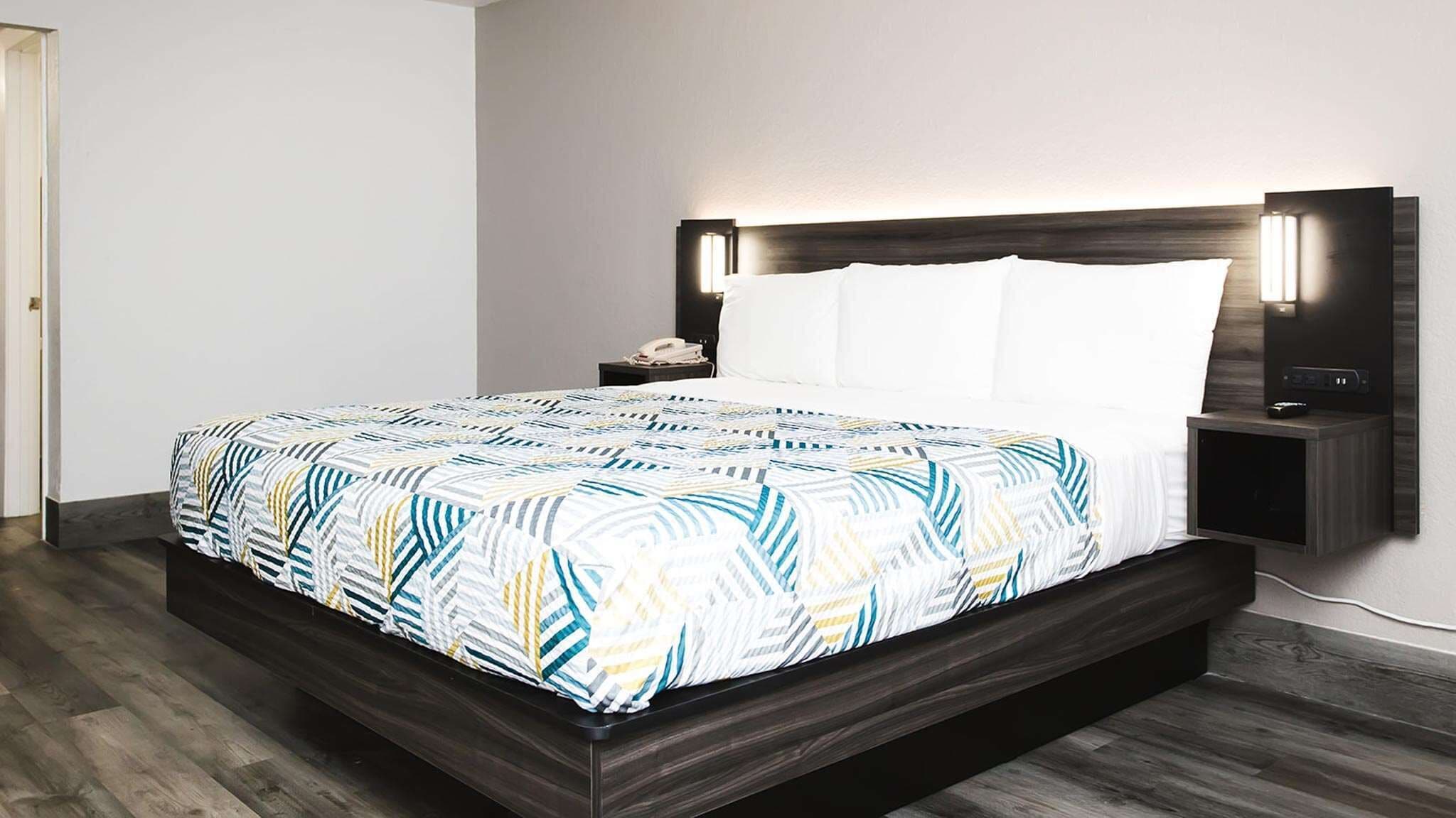 A modern hotel room with a large bed, a bedside table with a lamp, and a wall-mounted TV.