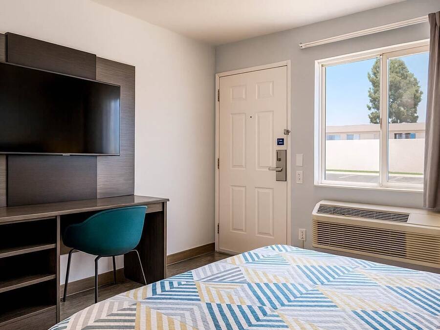 Habitación de hotel con una cama, un escritorio con silla, un televisor de pantalla plana en la pared, una puerta y una ventana con cortinas.
