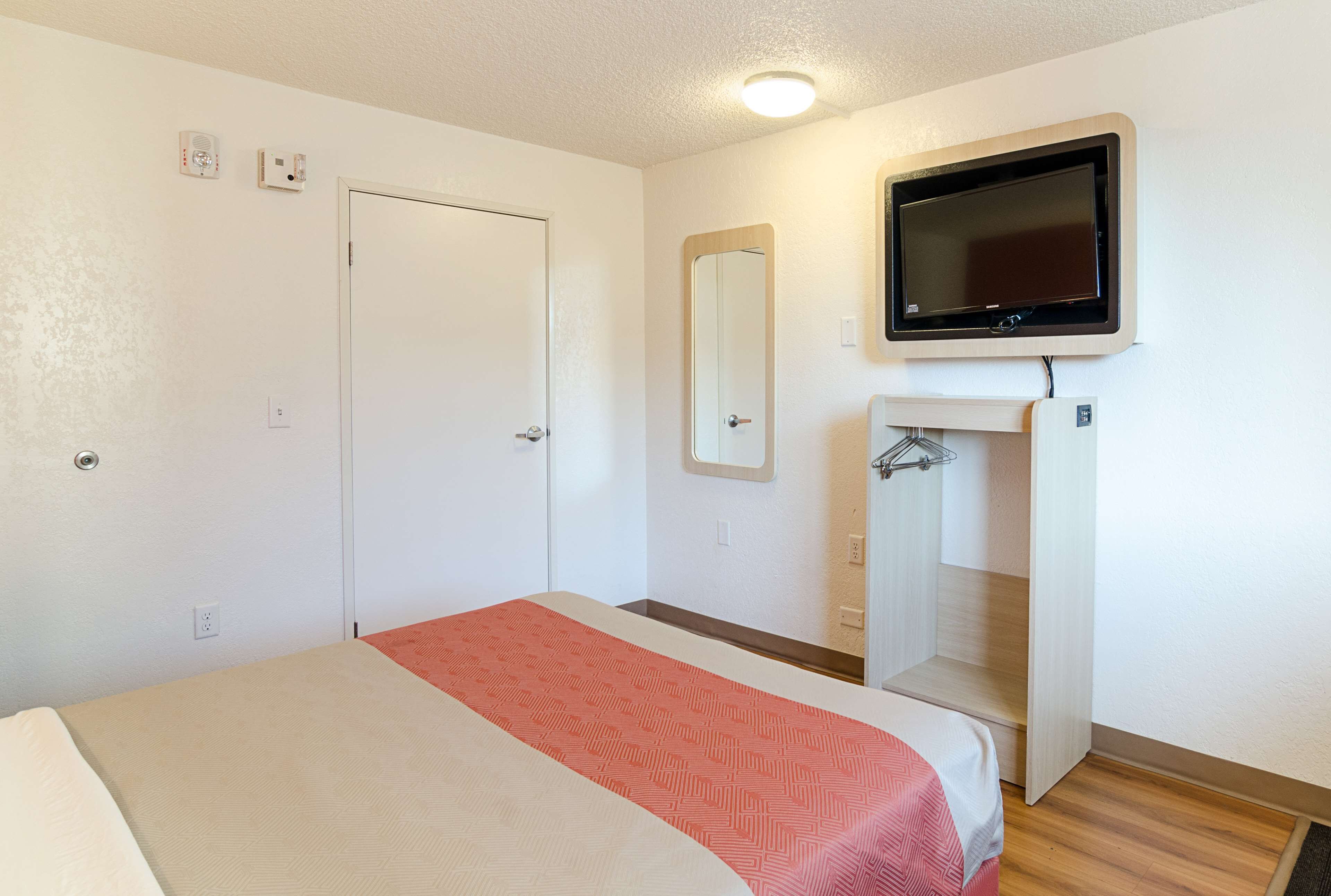 Una habitación de hotel con una cama, televisión montada en la pared, una puerta y un espejo.