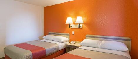 A hotel room with two beds, each with a pillow and a blanket. There is a nightstand with a phone between the beds. The room has orange walls and a white ceiling.